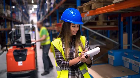 Portrait of female warehouse worker checking inventory in storage department while her coworker operating forklift in background.