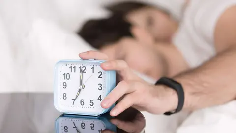 Young couple awaking after wake-up signal in the morning at home. Sleepy man stretching hand to alarm clock touching button to snooze or turn it off. Focus on hand with clock. Time to wake up